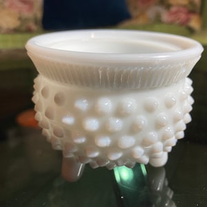 Puede incluir: Un plato de vidrio de leche blanco con un borde festoneado y una superficie texturizada y punteada. El plato tiene tres pequeños pies y es perfecto para guardar objetos pequeños.