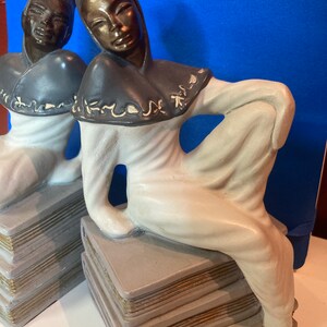 May include: Pair of Art Deco-style figurines. Each figurine depicts a seated figure in a flowing white robe and dark hood, resting on a stack of books. The faces and hoods have a bronze finish, and the books have gold-colored edges.