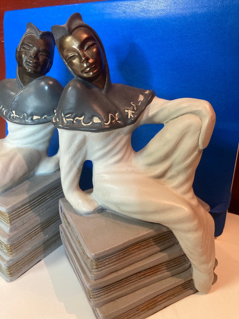 May include: Pair of Art Deco-style figurines. Each figurine depicts a seated figure in a flowing white robe and dark hood, resting on a stack of books. The faces and hoods have a bronze finish, and the books have gold-colored edges.