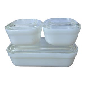 以下が含まれることがあります： ヴィンテージの白いガラス製食品保存容器のセット。セットには、2つの小さな正方形の容器と1つの長方形の容器が含まれており、すべて透明なガラスの蓋が付いています。容器は積み重ねられています。