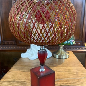 May include: A vintage table lamp with a woven, spherical shade in red, gold, and tan. The lamp has a red, cube-shaped base and a dark red, decorative stem. The lamp is on a wooden table.