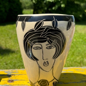 Könnte beinhalten: Eine Keramikvase mit einem schwarz-weißen Design eines Frauenkopfes. Die Vase hat einen cremefarbenen Hintergrund und schwarze Akzente.