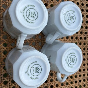 May include: Four white ceramic mugs with a green and white logo that reads "KAHLA German Democratic Republic" and a crown. The mugs are on a woven wicker surface.