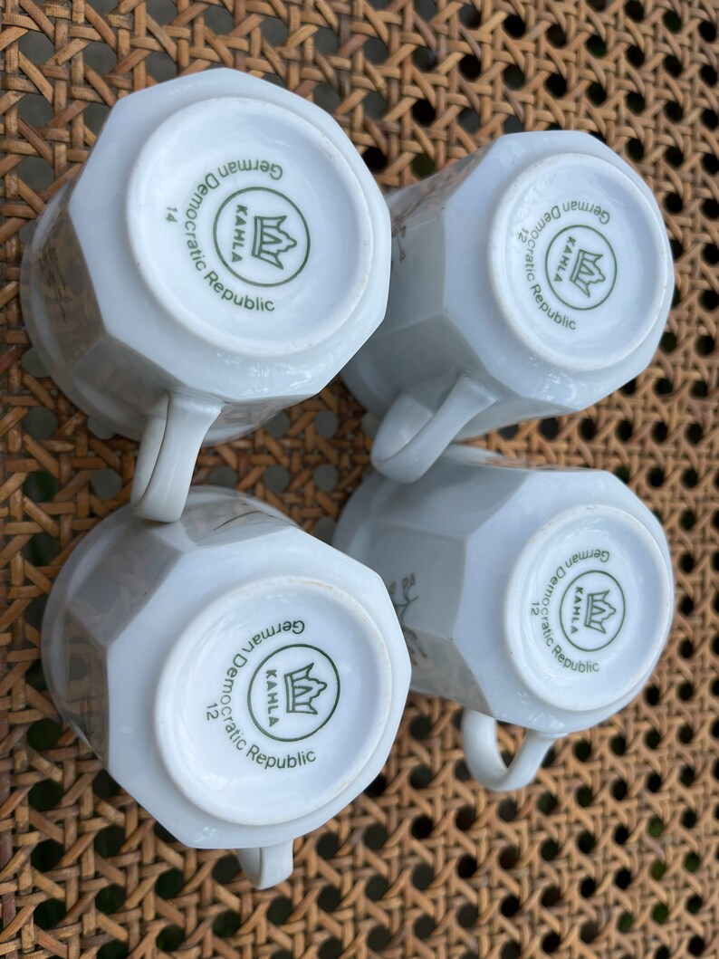 May include: Four white ceramic mugs with a green and white logo that reads "KAHLA German Democratic Republic" and a crown. The mugs are on a woven wicker surface.