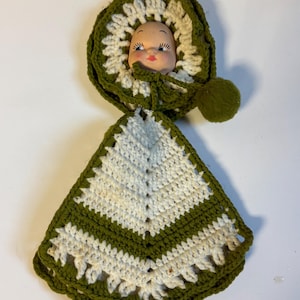 Puede incluir: Una muñeca vintage hecha a mano con un gorro y babero de ganchillo verde y blanco. La cara de la muñeca es visible, con rasgos pintados. El gorro tiene un pompón y una corbata. El babero es triangular con un borde festoneado.