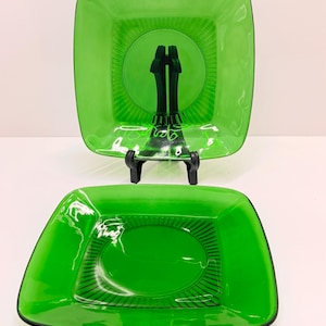Può includere: Due piatti da portata in vetro verde. Il piatto superiore è quadrato con un disegno circolare al centro ed è sostenuto da un supporto nero. Il piatto inferiore è rettangolare con un design ovale e bordi neri.