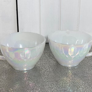 May include: Two white iridescent teacups with a textured design. The cups have a handle on the right side.