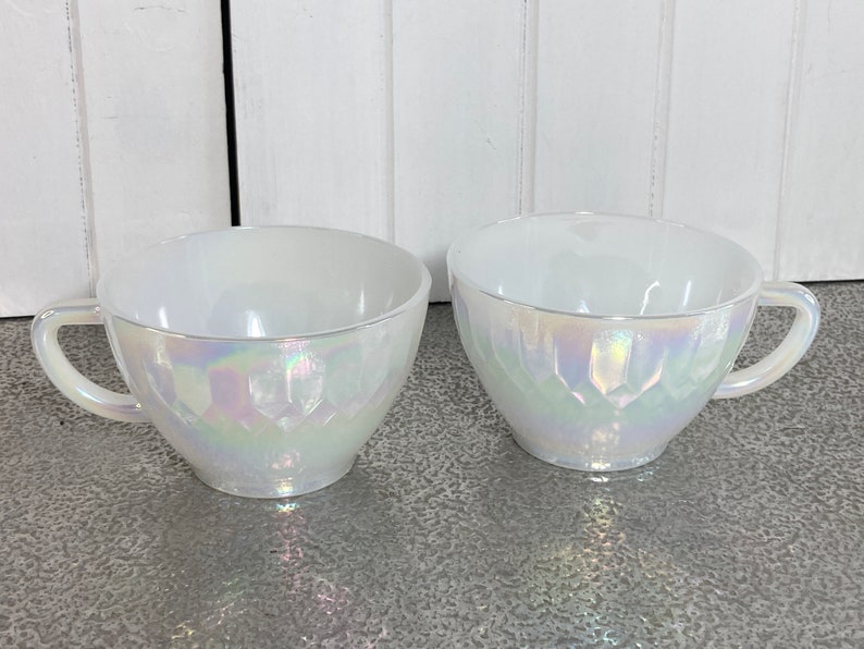 May include: Two white iridescent teacups with a textured design. The cups have a handle on the right side.