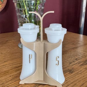 May include: A vintage salt and pepper shaker set. The white shakers have a beige plastic holder with a handle. The letters "P" and "S" are in gold. The shakers have white lids. A vase of flowers is in the background.
