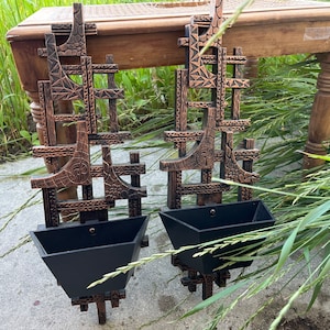 May include: Two copper-toned metal wall planters with geometric designs and black plastic pots. The planters are hanging on a wooden table.