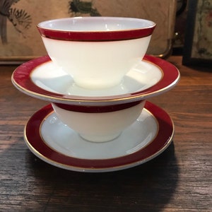 Può includere: Due piattini in ceramica bianca con un bordo bordeaux e un accento dorato. I piattini sono impilati uno sopra l'altro. Il piattino superiore ha una tazza in ceramica bianca con un bordo bordeaux e un accento dorato.