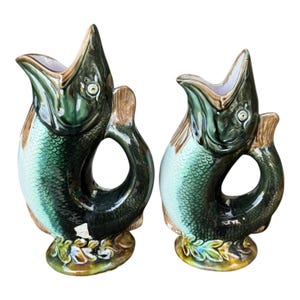 May include: Two ceramic fish-shaped jugs. The jugs have a green and blue scale pattern, with brown accents on the fins and a cream-coloured interior. The fish have open mouths and a handle. The base of the jugs have a floral design.