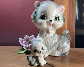 Op de afbeelding: Een paar keramische kattenbeeldjes. De grotere kat is grijs en wit met groene ogen en een roze neus. De kleinere kat is grijs en wit met groene ogen en een roze neus. De katten zitten op een houten oppervlak.