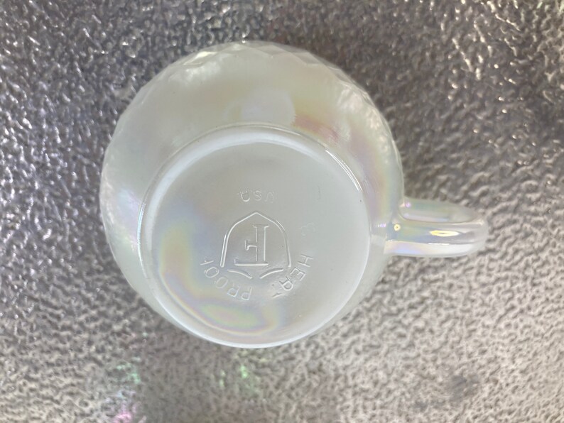 May include: White iridescent ceramic mug with a handle. The bottom of the mug has the text "HEIRLOOM PROOF" and a logo with the letter "E" inside a shield. The text "USA" is also visible.