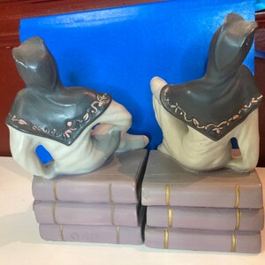 May include: A pair of ceramic bookends featuring figures in hooded cloaks, seated atop stacks of books. The cloaks are dark gray with pink and white floral trim. The books are lavender with gold accents.
