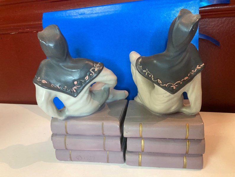 May include: A pair of ceramic bookends featuring figures in hooded cloaks, seated atop stacks of books. The cloaks are dark gray with pink and white floral trim. The books are lavender with gold accents.