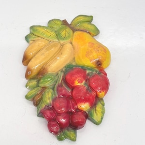 Peut inclure: Une décoration murale en céramique représentant un groupe de fruits, dont une poire, des bananes, des fraises et des cerises. Les fruits sont peints dans des couleurs réalistes, avec des feuilles et des tiges vertes.