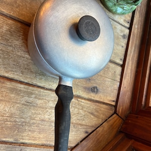 May include: A vintage aluminium cooking pot with a black handle and lid. The lid features a black knob with a circular design. The pot has a rounded shape and a long handle. A small metal ring is attached to the end of the handle.