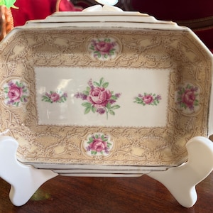 Op de afbeelding: Rechthoekige vintage serveerschaal met een crèmekleurige achtergrond en een bloemenrozenontwerp. De schaal heeft een decoratieve kantachtige rand en wordt ondersteund door een witte standaard. De rozen zijn roze en paars met groene bladeren.