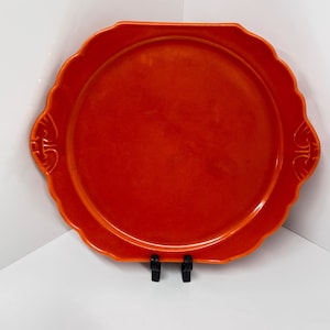 Peut inclure: Un plateau de service orange vif avec un bord festonné et des poignées décoratives. Le plateau rond a un bord légèrement surélevé et est soutenu par deux supports noirs. Le plateau est en matériau solide.