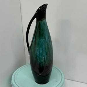 Puede incluir: Un jarrón alto y esbelto con asa, con acabado brillante. El jarrón hace la transición de una parte superior negra a un cuerpo verde y turquesa. El jarrón mide aproximadamente 30 cm de alto.
