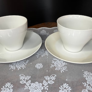 Puede incluir: Dos tazas de té y platillos de porcelana blanca sobre un mantel de encaje blanco. Las tazas tienen un diseño simple y elegante.