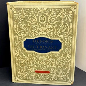 May include: An antique Oxford International Dictionary of the English Language. The book features a gold cover with elaborate scrollwork and a blue label with white text. The publisher's name is visible in red at the bottom.