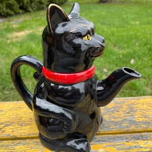 Può includere: Teiera in ceramica nera a forma di gatto con un colletto rosso. La teiera ha un design dipinto in oro sul viso del gatto e un beccuccio sul lato destro.