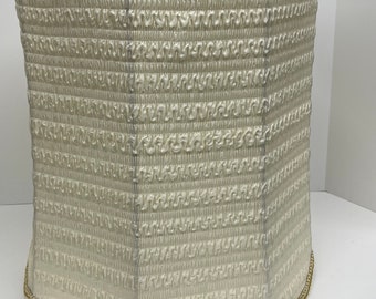 May include: Off-white lampshade with a textured, pleated design. The shade is trimmed with gold-colored decorative trim at the top and bottom edges. The lampshade has a cylindrical shape.