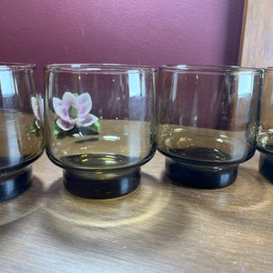 Può includere: Quattro bicchieri di vetro trasparente con una tinta marrone scuro e una base spessa. I bicchieri sono disposti in fila su una superficie di legno.