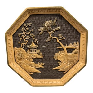 May include: An octagonal decorative tray with a gold-colored border and a dark brown center. The center features a detailed relief of a landscape with a pagoda, trees, and a boat. The tray's border has a repeating geometric pattern.