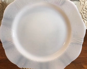 May include: A white milk glass plate with a scalloped edge. The plate is on a wooden surface and is being held up by a gold metal stand.