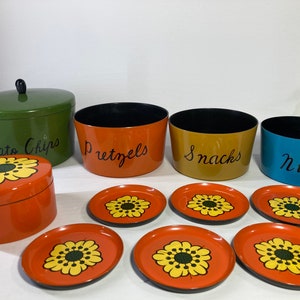 Op de afbeelding: Een verzameling vintage snackcontainers en onderzetters. De set bevat een groene container met het opschrift "Potato Chips", oranje containers met het opschrift "Pretzels", een gele container met het opschrift "Snacks" en een blauwe container met het opschrift "Nuts". Oranje onderzetters en een bijpassende doos hebben een geel bloemmotief.
