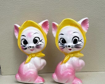 May include: Two ceramic cat ornaments. Each cat is pink with a yellow bonnet and bow. The cats have black whiskers and large eyes. The figurines are vintage-style and decorative.
