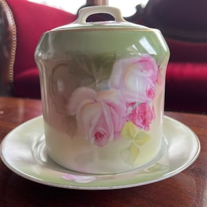 May include: A vintage porcelain sugar bowl with a lid and a matching saucer. The bowl and saucer are decorated with a floral design featuring pink roses and green leaves.