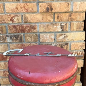 May include: A silver-toned metal emblem featuring the word Custom in a cursive script font. The emblem has a long, thin horizontal line extending from the letter C and ending after the letter m, with a metallic finish and a smooth surface.