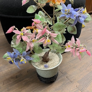 May include: A glass flower arrangement in a green ceramic pot with a black base. The flowers are pink, blue, and orange. The arrangement is on a wooden floor.