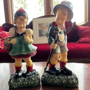Op de afbeelding: Twee vintage beeldjes van kinderen gekleed in traditionele Duitse kleding. Het meisje houdt een mand met bloemen vast en de jongen houdt een wandelstok vast. Beide beeldjes staan op een groene basis.