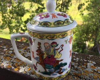 Puede incluir: Taza de té de porcelana blanca con tapa, con un diseño chino colorido de un hombre montando un ciervo. La taza tiene un asa y un patrón floral alrededor del borde.