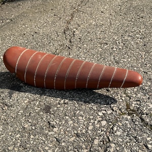 Op de afbeelding: Een bruin leren fietszadel met witte stiksels. Het zadel is lang en smal, met een licht gebogen vorm.