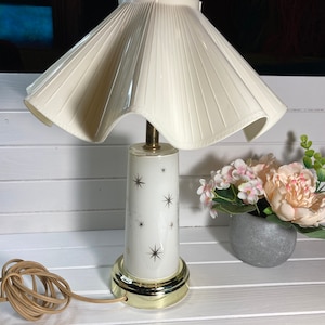 May include: A white table lamp with a pleated white shade and a white base with a gold trim. The base has a star pattern. The lamp is on a white surface with a small potted plant with pink flowers.