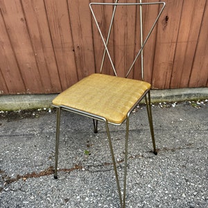 Op de afbeelding: Een mid-century modern stoel met een metalen frame en een gestoffeerde zitting in mosterdgeel.