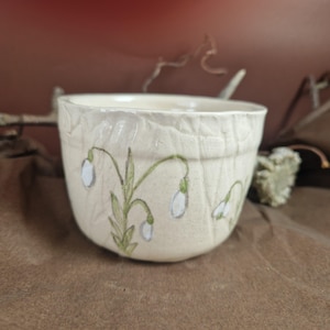 May include: A white ceramic bowl with a textured surface and a design of white snowdrops with green stems.