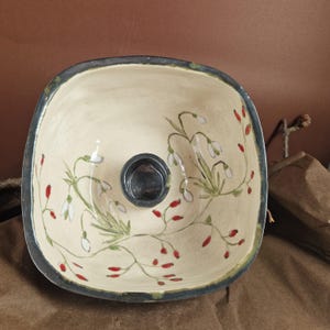 Op de afbeelding: Een vierkante keramische kom met een witte glazuur en een zwarte rand. De kom is versierd met handgeschilderde witte sneeuwklokjes en rode bessen.