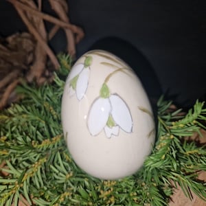 Może przedstawiać: Kremowy, dekoracyjny przedmiot w kształcie jaja z ręcznie malowanymi białymi kwiatami przebiśniegów z zielonymi akcentami. Jajko otoczone jest zielonymi liśćmi, tworząc naturalną, wiosenną estetykę.