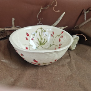 May include: A white ceramic bowl with a floral design. The bowl is decorated with red berries and white flowers. The bowl is handmade and has a rustic look.