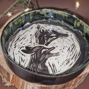 Könnte beinhalten: Eine schwarze und weiße Keramikschüssel mit grünem Glase am Rand. Die Schüssel zeigt ein stilisiertes Bild von zwei Füchsen, die sich gegenüberstehen.