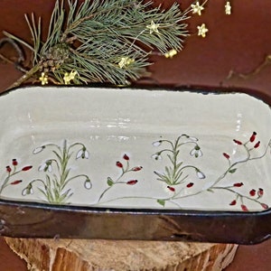 May include: A rectangular ceramic baking dish with a white glaze and a black rim. The dish is decorated with a hand-painted design of white snowdrops and red berries on green stems.