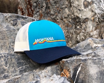 Puede incluir: Una gorra de camionero azul y blanca con una visera azul marino. La gorra tiene un gráfico de montaña arcoíris y la palabra "MONTANA" bordada en la parte delantera.