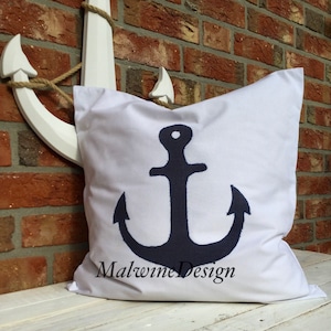 Könnte beinhalten: Weißes quadratisches Kissen mit einem marineblauen Ankerdesign und dem Text "Malwine Design". Im Hintergrund befindet sich ein weißer Anker, dahinter eine Backsteinmauer.